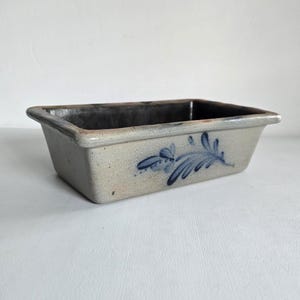 May include: A rectangular stoneware dish with a light gray exterior and a dark interior. The dish features a blue floral design on the side. The stoneware dish is a vintage piece.