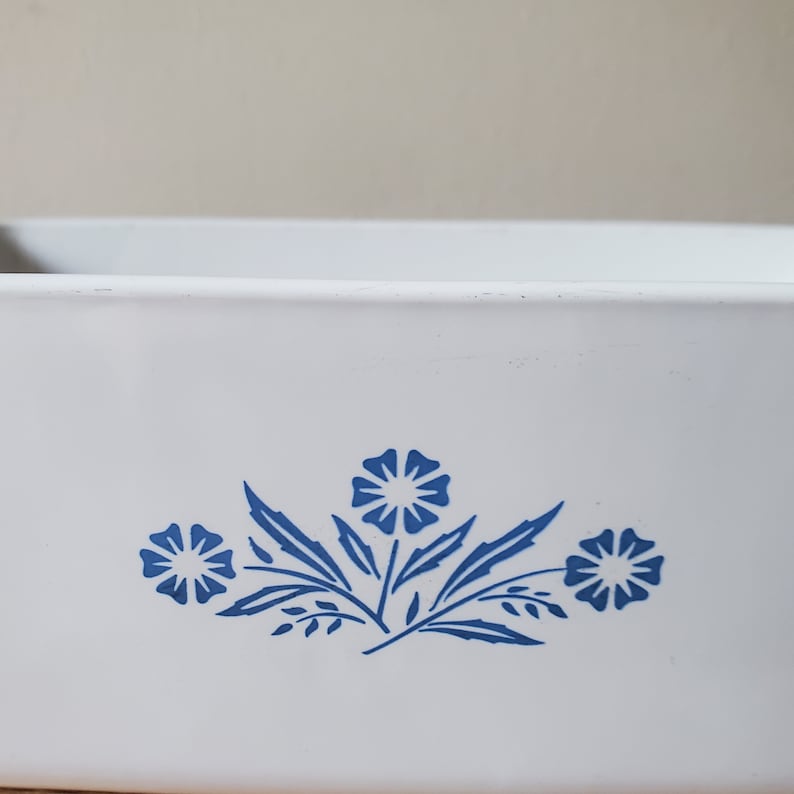 Puede incluir: Plato para hornear de cer&aacute;mica blanca con un dise&ntilde;o floral azul. El dise&ntilde;o presenta tres flores estilizadas con hojas.