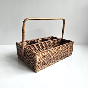 May include: A rectangular woven rattan basket featuring a natural brown finish, a high arched handle, and an interior divided into three separate compartments by two crosswise woven dividers, creating an organized storage structure.