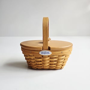 May include: A light brown woven basket with a wooden lid and handle. The basket has a rounded shape and a hinged lid. The handle is arched and attached to the lid. A small blue and white label on the front reads "CENTURY".