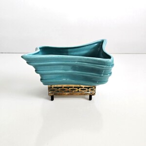 May include: A turquoise ceramic planter with a wavy, textured design. The planter sits on a decorative gold-colored metal stand with a geometric pattern. The planter's shape is asymmetrical, with a flared rim.