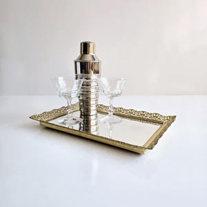 May include: A silver cocktail shaker and two clear coupe glasses sit on a gold-toned rectangular mirrored tray with a decorative edge. The tray is on a white surface, creating a classic barware arrangement.