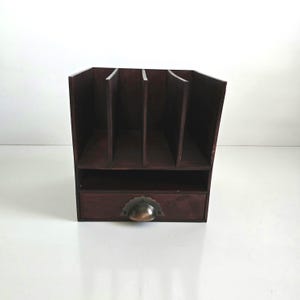 May include: A dark brown wooden desk organizer with multiple vertical slots and a small drawer. The drawer has a bronze-colored, half-moon shaped handle. The organizer is designed to hold papers, files, or other office supplies.