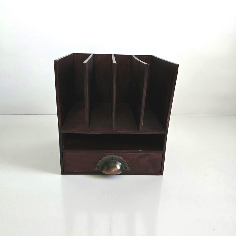 May include: A dark brown wooden desk organizer with multiple vertical slots and a small drawer. The drawer has a bronze-colored, half-moon shaped handle. The organizer is designed to hold papers, files, or other office supplies.