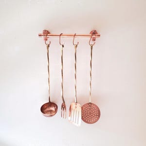 May include: Copper kitchen utensil set hanging on a copper bar. The set includes a ladle, fork, spatula, and slotted spoon. The utensils have twisted handles and are suspended from hooks on the bar.