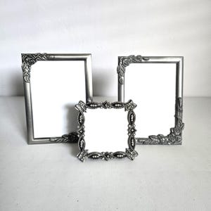 May include: Three silver picture frames. Two rectangular frames feature floral corner accents. A smaller square frame has a decorative beaded border. All frames have white interiors.