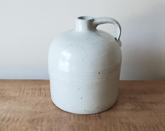 May include: A white ceramic jug with a handle. The jug has a rounded body and a slightly flared lip.