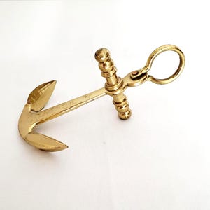 May include: A brass anchor-shaped object with a ring handle. The anchor has a classic design with a curved fluke and a central stock. The brass has a warm, golden hue, suggesting an antique or vintage piece.