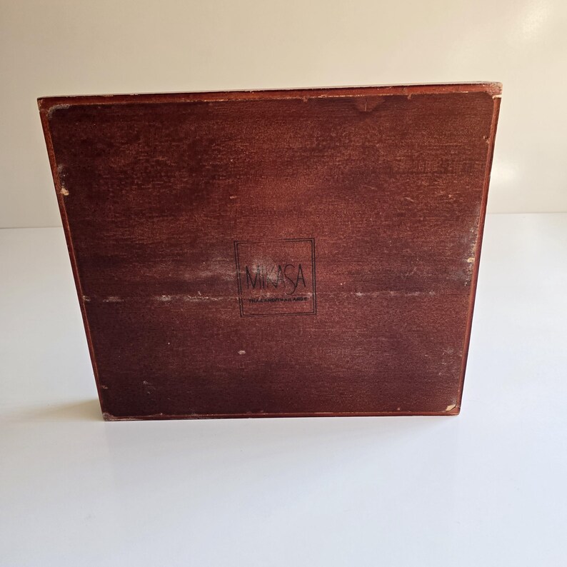 May include: A dark brown wooden box with a rectangular shape and angled sides. The box displays the "MIKASA" logo within a rectangular frame. The box is set against a white background.