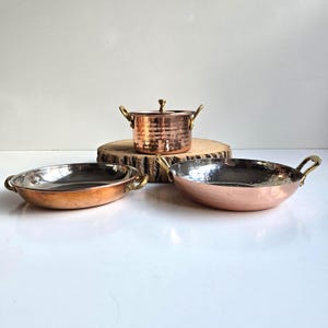 May include: A set of three copper cookware pieces including a lidded pot and two shallow pans. Each item features a hammered exterior finish, polished metallic interiors, and curved brass handles attached to the sides of the vessels.