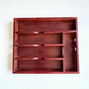 May include: A dark red wooden organiser with multiple compartments. It features three shelves with curved cutouts and a vertical section with two circular holes. The wood grain is visible throughout the piece.
