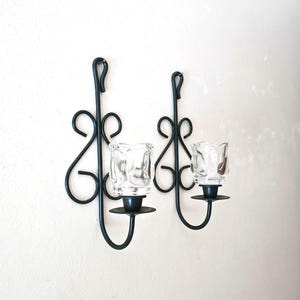 May include: A pair of black metal wall sconces featuring scrollwork designs and clear glass candle holders. Each sconce has a vertical frame with curved metal accents and a circular base to support the removable glass cup for a candle.