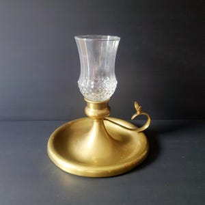 May include: A vintage brass candle holder with a clear glass cup. The candle holder has a round base with a small handle and a decorative design on the glass cup. The brass has a warm, golden tone.
