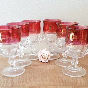May include: Set of six clear glass stemmed wine goblets with a ruby red iridescent finish. The goblets have a textured, knobby design and are arranged on a wooden surface.