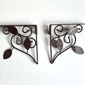 May include: A pair of dark brown metal shelf brackets featuring a right-angle frame with decorative scrollwork and attached leaf-shaped accents. Each bracket has a flat top edge and a vertical side edge, with metal vines and leaves integrated.