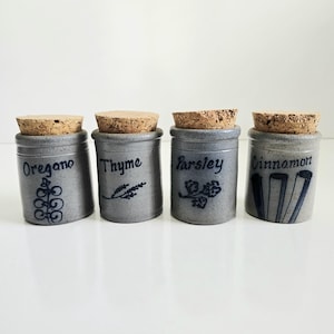 May include: Four gray ceramic spice jars with cork lids. Each jar is labeled with a different spice: oregano, thyme, parsley, and cinnamon. The labels are in a dark blue script with decorative illustrations.