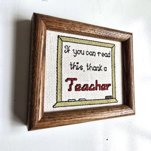 May include: Cross-stitch artwork in a wooden frame. The artwork features the text "If you can read this, thank a Teacher" in black and red thread on a white background, framed by a yellow border.