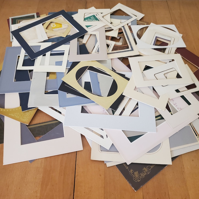 May include: A pile of various sized and colored picture frames, some with white mats, some with black mats, and some with gold mats. The frames are made of cardboard and are in various stages of wear and tear.