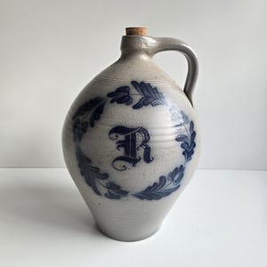 May include: A light gray stoneware jug with a single handle and a cork stopper. The front features a dark blue hand-painted letter R centered within a circular wreath of stylized leaves. The ceramic surface has a speckled texture throughout.