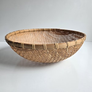 May include: A round, shallow bowl-shaped basket crafted from light brown woven bamboo strips. The basket features a fine, open-weave lattice pattern throughout the body and a reinforced, smooth bamboo rim secured with thin, tightly wrapped strips.