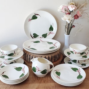 May include: A set of vintage white ceramic dishes with a green leaf and red berry design. The set includes a creamer, teacups, saucers, and dinner plates.