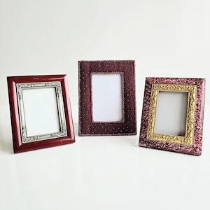 May include: Three decorative picture frames in varying styles. The frame on the left is a dark red wood with a silver inner frame. The center frame is a deep burgundy fabric with small beaded accents. The frame on the right has a gold inner frame and a textured, patterned exterior.