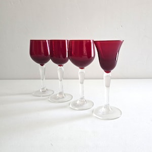 May include: Four ruby red wine glasses with clear stems and bases are arranged in a row. The glasses have a classic design with a rounded bowl and a slender stem. The background is a plain white surface.