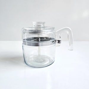 May include: A clear glass coffee percolator with a glass lid and handle. The percolator has a silver-colored band around the middle and a clear glass handle. The coffee maker is sitting on a white surface.