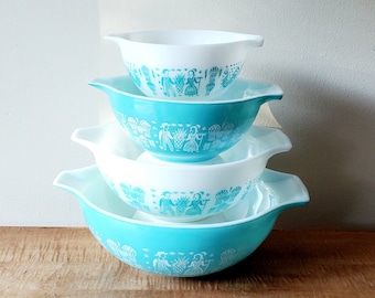 May include: A set of four nesting bowls in a turquoise blue colour with a white design of people in traditional clothing. The bowls are made of ceramic and have a glossy finish.