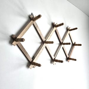 May include: An expandable wooden wall rack featuring a diamond-shaped lattice design with ten cylindrical pegs. The light brown wood has a natural grain finish, and the structure is mounted horizontally with metal hardware visible at the top.