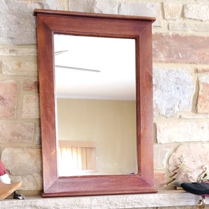 May include: Rectangular wooden framed mirror with a rich, dark brown finish. The mirror is mounted on a stone wall. The frame has a simple, classic design, suitable for various interior styles. The mirror reflects the room.