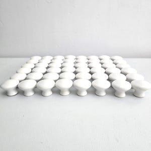 May include: A collection of white ceramic cabinet knobs arranged in rows. Each knob has a rounded top and a short, cylindrical base. The knobs are evenly spaced and appear to be new.