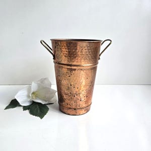 May include: A tall, tapered copper bucket featuring a hammered texture on the upper section and two side handles. The metal surface displays a weathered, mottled patina with dark spots throughout. A single white flower with green leaves sits nearby.