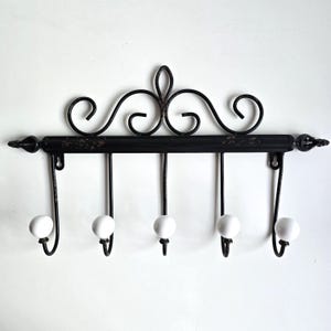 May include: Black metal wall-mounted coat rack with five hooks. The rack features decorative scrollwork and white ceramic ball-shaped hook ends. The design is a classic, vintage style, suitable for entryways or bedrooms.