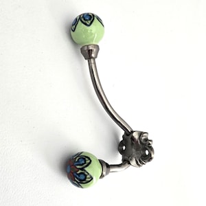 May include: A wall-mounted metal hook featuring two curved arms, each ending in a spherical ceramic knob. The knobs are light green with a painted floral pattern in blue, red, and black. The metal base has a dark, weathered finish.