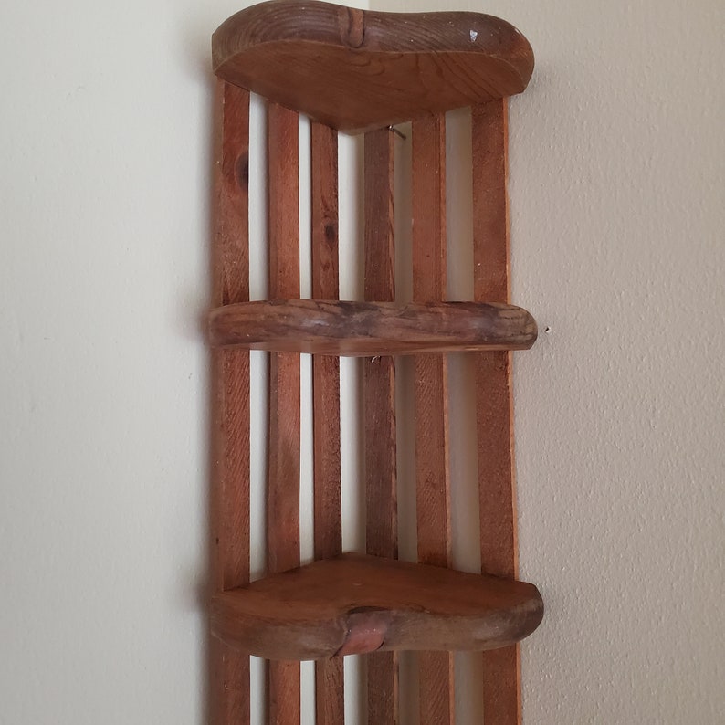 May include: A wooden corner shelf with three shelves. The shelves are made of wood slats and have a rounded edge. The shelf is a light brown color.