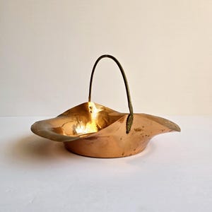 May include: A copper-colored metal basket with a curved handle. The basket has a wide, shallow bowl shape with a gently undulating rim. The handle is a darker color, possibly bronze or brass.