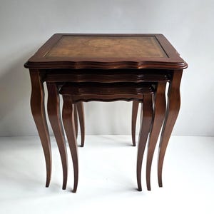 May include: A set of three nested tables crafted from dark brown wood, featuring curved cabriole legs and rectangular tops. The largest table has a central inlay panel with a mottled tan and brown finish, while all tables have scalloped edges.