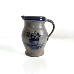May include: A gray ceramic pitcher with a dark blue handle and rim. The pitcher features a hand-painted design of a potted plant in dark blue. The pitcher has a spout for pouring and a curved handle.