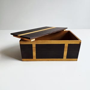 May include: A rectangular wooden box with a hinged lid, partially open. The box features a dark brown exterior with light brown accents and a light brown interior. The lid has a dark brown finish with a light brown stripe.