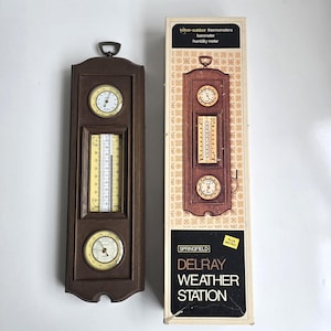 May include: A brown wall-mounted weather station featuring a thermometer, barometer, and humidity meter with a yellow background. The original packaging box is included, labeled Springfield Delray Weather Station, displaying the same device.