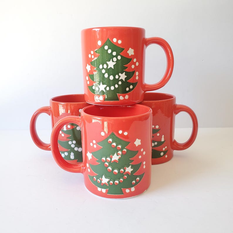 May include: Four red ceramic mugs stacked, each featuring a green Christmas tree design with white star and dot ornaments. The mugs have a curved handle and a glossy finish, perfect for holiday beverages.