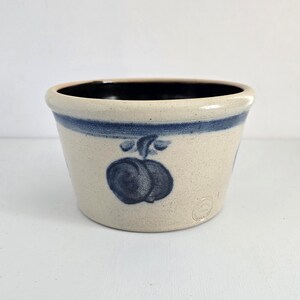 May include: A small, round, stoneware bowl with a cream-colored exterior and a dark blue interior. The bowl features a dark blue band around the top and a dark blue fruit design on the front. The bowl is likely used for food or decoration.