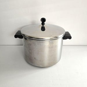 May include: A stainless steel cooking pot with a lid and black handles. The pot has a round body and a shiny, metallic finish. The lid has a black knob. The pot is designed for cooking and food preparation.