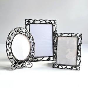 May include: A set of three silver-toned metal picture frames featuring an intricate, raised vine and leaf motif. The collection includes one oval frame and two rectangular frames, all with a dark, weathered finish and openwork borders.
