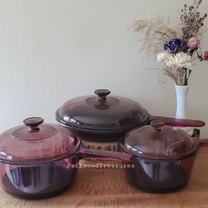 May include: Three purple glass cookware lids with handles. The lids are on a wooden surface.