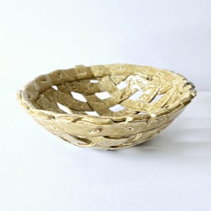 May include: A woven ceramic bowl with a light beige glaze. The bowl has a basket-like design with an open weave pattern. The rim and base are rounded, and the bowl is set against a white background.