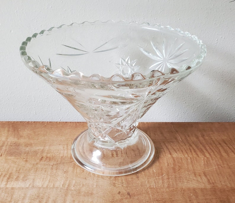May include: A clear glass bowl with a starburst pattern. The bowl has a scalloped edge and sits on a pedestal base.