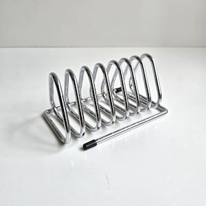 May include: A chrome-finished metal toast rack featuring seven triangular slots for holding slices of bread. The structure is composed of a rectangular base frame with a single black rubber tip on one end of the wire frame for stability.