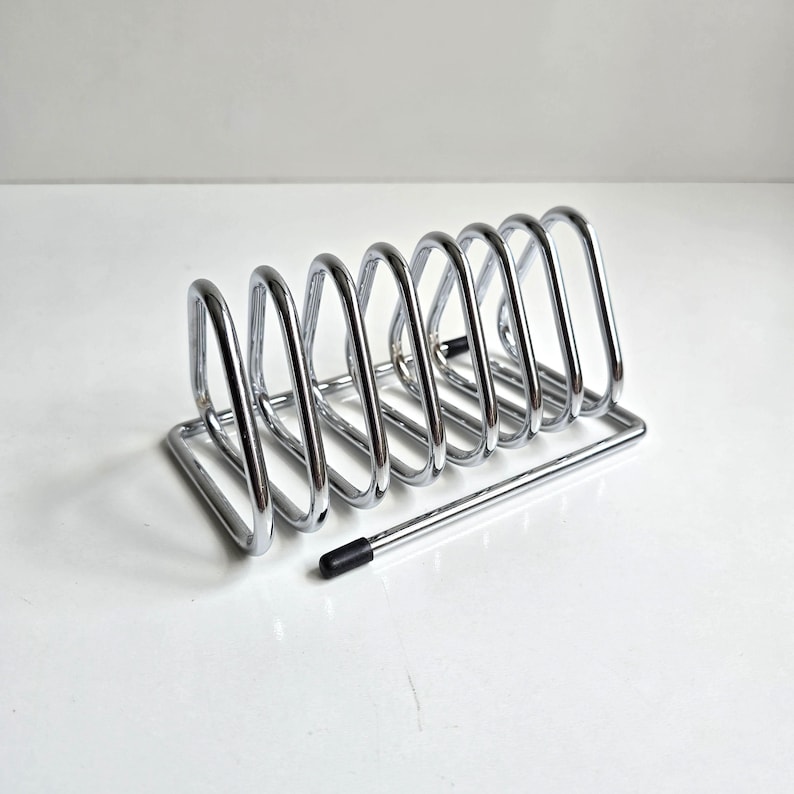May include: A chrome-finished metal toast rack featuring seven triangular slots for holding slices of bread. The structure is composed of a rectangular base frame with a single black rubber tip on one end of the wire frame for stability.